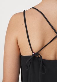 Gros plan sur le dos d'une femme portant un haut noir à fines bretelles croisées, nouées en un nœud au centre de son haut du dos.