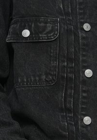 Μαύρο τζάκετ από τζιν με ασημένια κουμπιά, τσέπη στο στήθος και υφή επιφάνειας. Ο σχεδιασμός περιλαμβάνει ορατές ραφές και κλασική γραμμή.