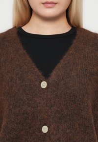 Gros plan d'une personne portant un cardigan marron duveteux avec deux boutons beiges sur un haut côtelé noir, ne montrant que le cou et le menton.