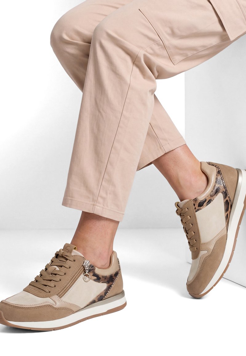 Personne portant des baskets beiges et marron avec des détails imprimé léopard, associées à un pantalon beige clair court, assise avec un pied levé.