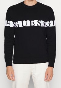 Homme portant une chemise noire à manches longues avec un texte blanc en gras déformé sur la poitrine et les bras supérieurs, assortie à un pantalon de couleur claire.