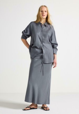 Ung kvinde med langt blondt hår iført en løs grå skjorte med knapper, matchende langt skørt og sorte sandaler mod en ensartet baggrund.