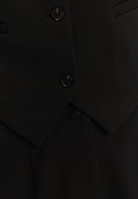 Nahaufnahme eines schwarzen, maßgeschneiderten Blazers mit zwei sichtbaren schwarzen Knöpfen und einer Taschendetail auf strukturiertem Stoff.