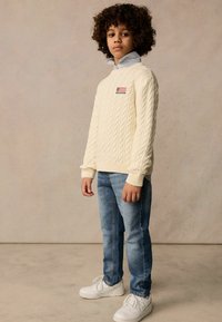 Cremfarbener Kabelstrickpullover mit einem Patch der amerikanischen Flagge, leicht blaues Hemd darunter, blaue Jeans und weiße Sneakers.