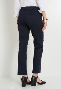 Persona vestida con pantalones de vestir azul marino, camisa blanca con mangas remangadas, zapatos de tacón negros y una pulsera de oro en la muñeca, de pie contra un fondo blanco.