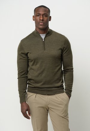 Suéter de punto verde oliva con cuello de cremallera de un cuarto, mangas largas y puños acanalados. Usado sobre una camisa gris clara, combinado con pantalones beige.