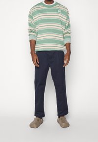 Pruhovaná zeleno-béžová mikina s logem, kombinovaná s tmavě modrými kalhotami a béžovými nazouvacími botami. Volný střih a kulatý výstřih.