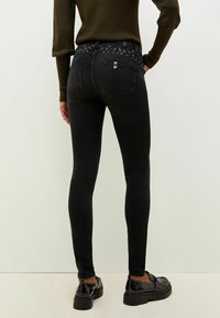 Vaqueros negros ajustados con un diseño ceñido, que cuentan con una cinturilla estampada y dos bolsillos traseros. Combinados con zapatos negros brillantes.