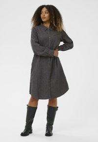 Sötét szürke farmeringruha, gallérral, gombos elülső résszel és hosszú ujjal. Térdépig érő fekete bőrcipővel viselve.