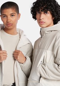 Beige Kapuzenjacken mit Reißverschluss und strukturierten Innenfuttern. Das Modell links hält die Jacke, während das Modell rechts eine glatte Stoffoberfläche hat.