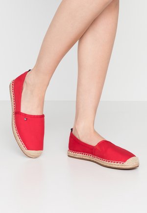 Personne portant des espadrilles rouges avec des semelles tissées beiges, se tenant sur un sol blanc devant un fond blanc uni.