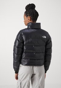 Črn puhovko s visokim ovratnikom, ki ima vodoravno prešite dele in znak na hrbtu. Tkanina se zdi sijoča in toplotno izolirana.