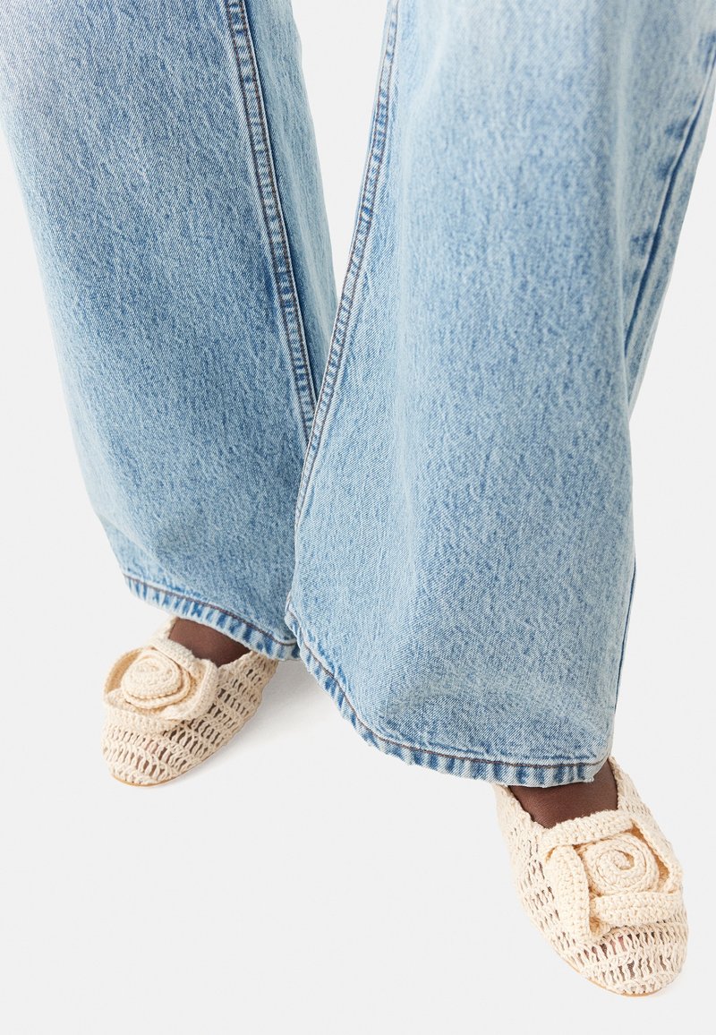 Zapatillas de croché beige con un diseño texturizado y un patrón de rosas en la parte frontal, llevadas con jeans de mezclilla de pierna ancha en azul claro.
