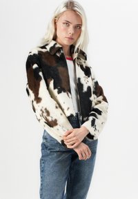 Jachetă din blană sintetică cu un imprimeu de vacă maro și alb, având fermoar frontal, guler clasic și mâneci lungi. Pur și simplu cu blugi de denim.