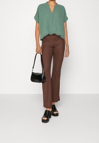 Haut vert à manches courtes, pantalon évasé côtelé marron, sandales à talons noires, et petit sac à main noir dans un décor minimaliste.