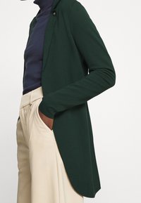 Dark green cardigan with a soft texture, long sleeves, open front, and a subtle clasp. Paired with a fitted navy top and beige trousers.