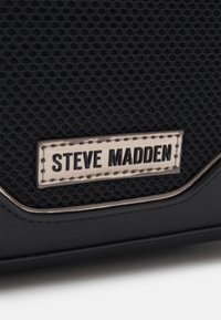Juodas tekstūruotas audinys ir odinis rankinukas, kuriame yra sidabrinė plokštelė su įspaudu "Steve Madden", pabrėžianti elegantišką ir modernų dizainą.