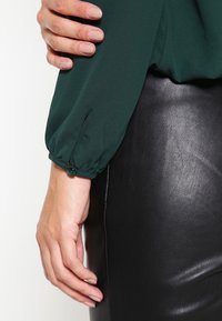 Blusa verde con maniche a sbuffo e dettagli con polsini elasticizzati, abbinata a una gonna nera in pelle. Mano appoggiata sul tessuto, mostrando una trama morbida.