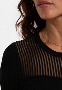 Haut noir avec texture côtelée et panneau transparent rayé, col rond, et un fin collier en or. Présente une coupe slim.