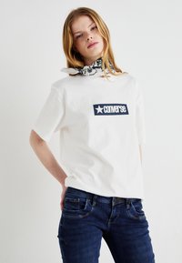 Biely tričko s modrým obdĺžnikovým logom "Converse" s hviezdou. Nosené s tmavomodrými džínsami a čierno-bielym vzorovaným šálom.
