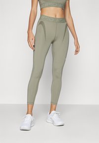 Olivenfargede leggings med teksturert midjebånd, slim passform og buede sømmer. Kombinert med hvite treningssko.