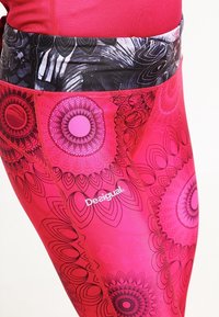 Rosa yogabyxor med intrikata blommönster i mörkare rosa nyanser. Midjebandet har en kontrasterande svart och vit design. Logotyp synlig.