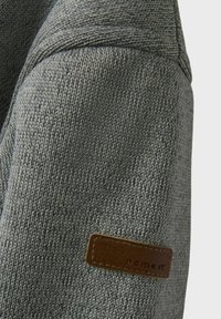 Un pull en tricot gris avec une surface texturée, présentant une étiquette en cuir marron sur la manche, mettant en avant un design moderne.