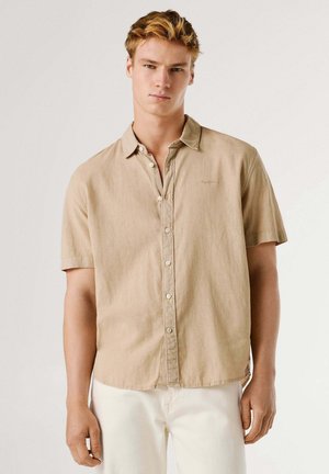 Giovane uomo con capelli biondi corti che indossa una camicia beige a maniche corte con bottoni e pantaloni bianchi, in piedi davanti a uno sfondo semplice.