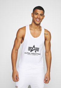 Bílé tílko s černým logem s ostrým designem a nápisem "ALPHA INDUSTRIES" pod ním. Vyrobeno z hladké látky. Jednoduchý střih.