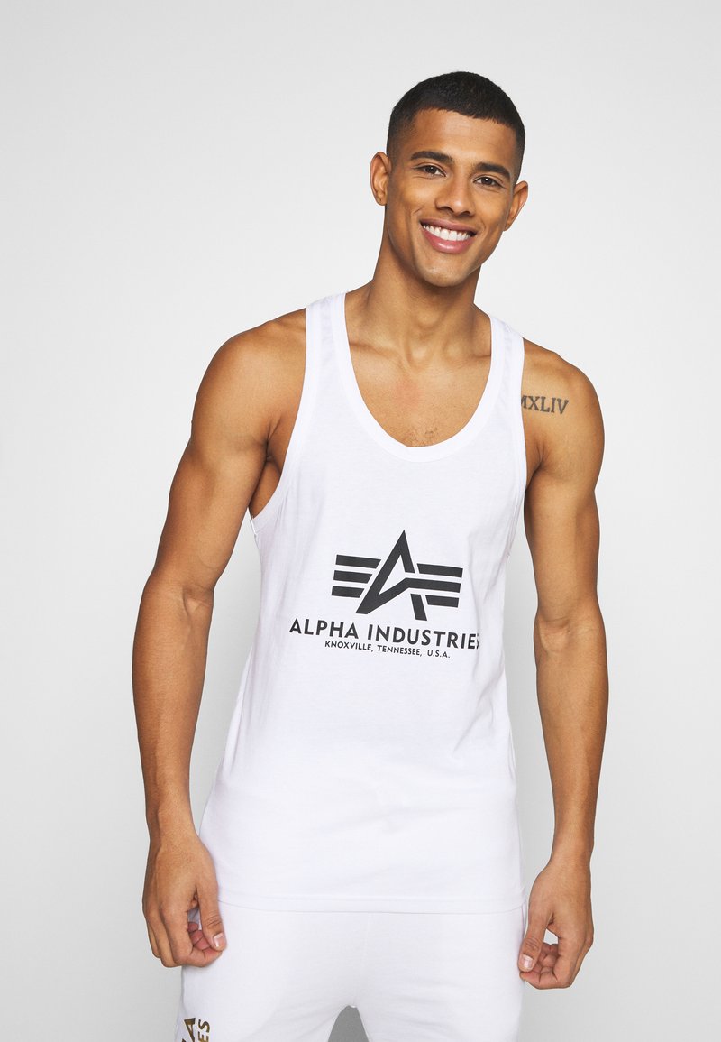 Bílé tílko s černým logem s ostrým designem a nápisem "ALPHA INDUSTRIES" pod ním. Vyrobeno z hladké látky. Jednoduchý střih.