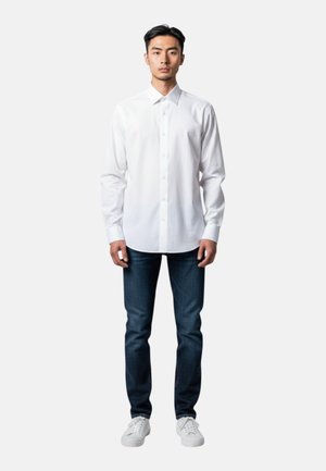 Uomo in piedi di fronte, indossa una camicia bianca con bottoni, jeans blu scuro e scarpe da ginnastica bianche su uno sfondo chiaro e uniforme.