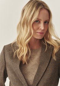 Rjava houndstooth blazer z vrhunskimi reverji, v kombinaciji s taupe puloverjem. Gladka tekstura tkanine, oprijet kroj in čiste linije.