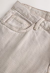 Jean beige avec un subtil motif en chevron, équipé d'une ceinture ajustée et de poches classiques. Le tissu présente une finition lisse et texturée.
