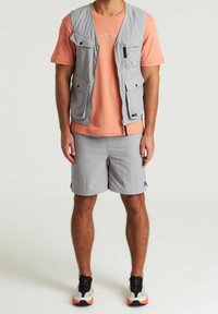 Gilet grigio chiaro con molte tasche sopra una camicia pesca a maniche corte; abbinato a pantaloncini grigio chiaro e sneaker nere con dettagli arancioni.
