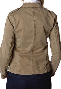 Femme portant une veste cintrée marron clair avec des poignets boutonnés, des détails de couture visibles et un jean bleu foncé, vue de dos.