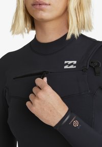 Nahaufnahme einer Person in einem schwarzen Neoprenanzug, die eine Brusttasche öffnet, wobei das Logo und die Größenmarkierung 3/2 Furnace auf dem Ärmel zu sehen sind.