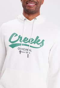 Sudadera blanca con un logo verde de "Creeks" y el texto "Original desde 1944". Cuenta con una capucha con cordones y un bolsillo canguro delantero.