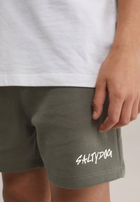 Olīvu zaļi šorti ar teksturētu audumu, ar baltu "SALTYDOG" uzrakstu apakšējā daļā un baltu T-kreklu virsū.