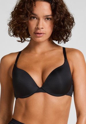 Femme aux cheveux bouclés portant un soutien-gorge noir rembourré, faisant face à l’avant sur un fond gris uni.