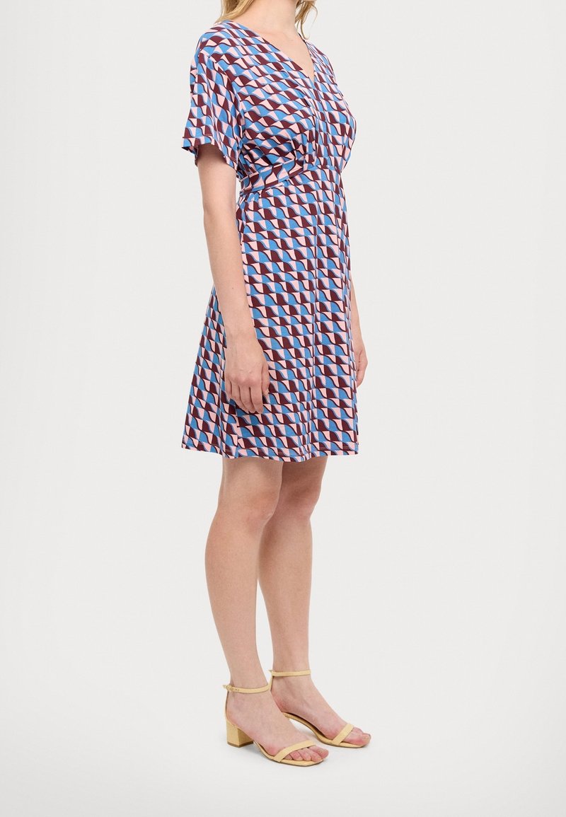 Femme portant une robe à motif géométrique longueur genou avec manches courtes et des sandales jaunes à talons blocs ouverts, debout de côté.