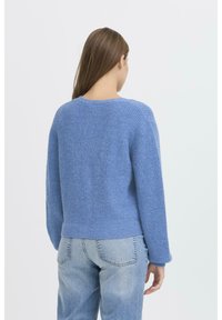 Maglione lavorato a maglia blu con finitura testurizzata, scollo rotondo e maniche lunghe, abbinato a jeans azzurri, visto da dietro.