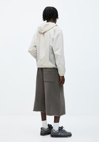 Man med ljusbeige hooded jacka, vida grå cropped-byxor med sidficka, vita strumpor och mörkgrå sneakers, stående vänd bort.