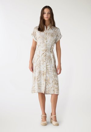 Donna in piedi con un vestito midi camicia beige e bianco a motivo di foglie di palma, maniche corte e sandali zeppa beige.