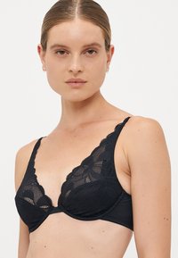 Vrouw met lichte huid en haar naar achteren vastgebonden draagt een zwarte kanten bh met gegolfde randen tegen een effen witte achtergrond.