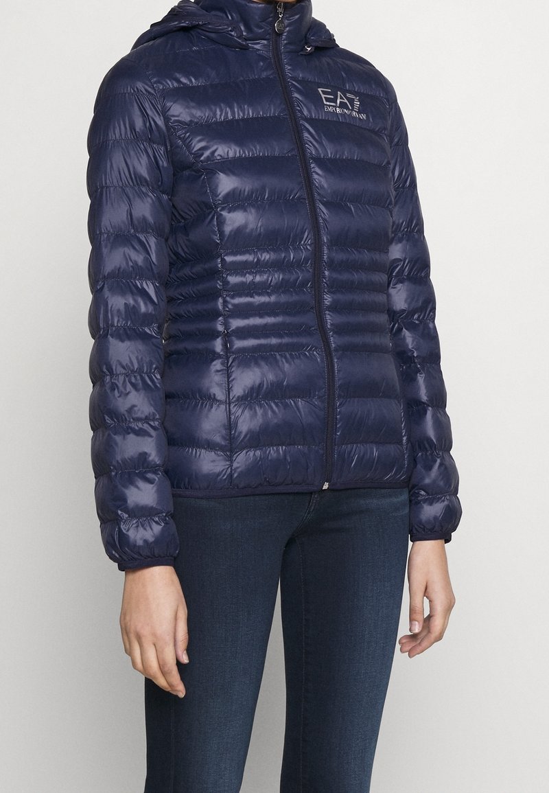 Chaqueta tipo puffer de color azul marino con costuras horizontales, cierre de cremallera y capucha. El material es brillante, ligero, con puños y dobladillo elásticos. Detalle del logo en el pecho.