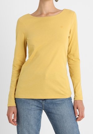 Kvinna i senapsgul långärmad tröja med rund halsringning och ljusblå denimjeans, stående mot en enfärgad bakgrund.