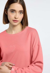 Koraalroze langepof top met een ronde halslijn, voorzien van een geborduurd hartdetail en een subtiel patroon op de manchetten. Zachte stoftextuur.