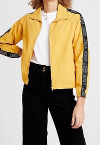 Żółta kurtka zapinana na zamek błyskawiczny z czarnymi akcentami logo na rękawach; lekki materiał, stójka i luźny krój.