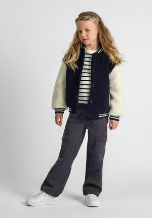 Sort og creme varsity jakke med blød tekstur, stribet hvid og sort sweater, grå cargo bukser og hvide sneakers.