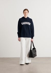 Sweatshirt azul-marinho com letras brancas "BARBOUR", combinado com calças brancas de perna larga e uma mala preta. Texturas suaves e design simples.